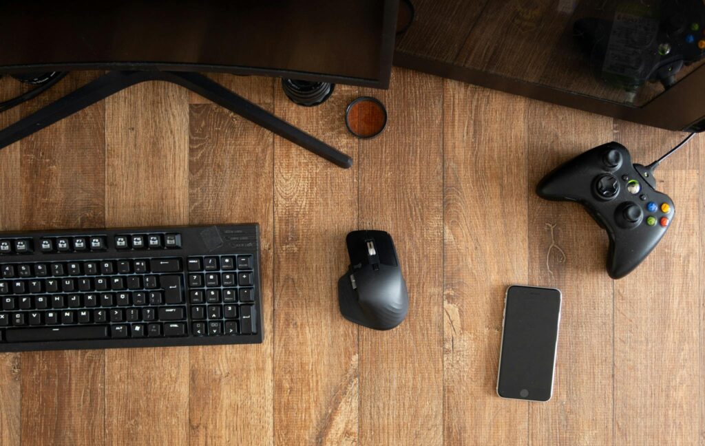 Top view of black gamepad with mouse and keyboard on wooden desk with monitor in apartment in daytime